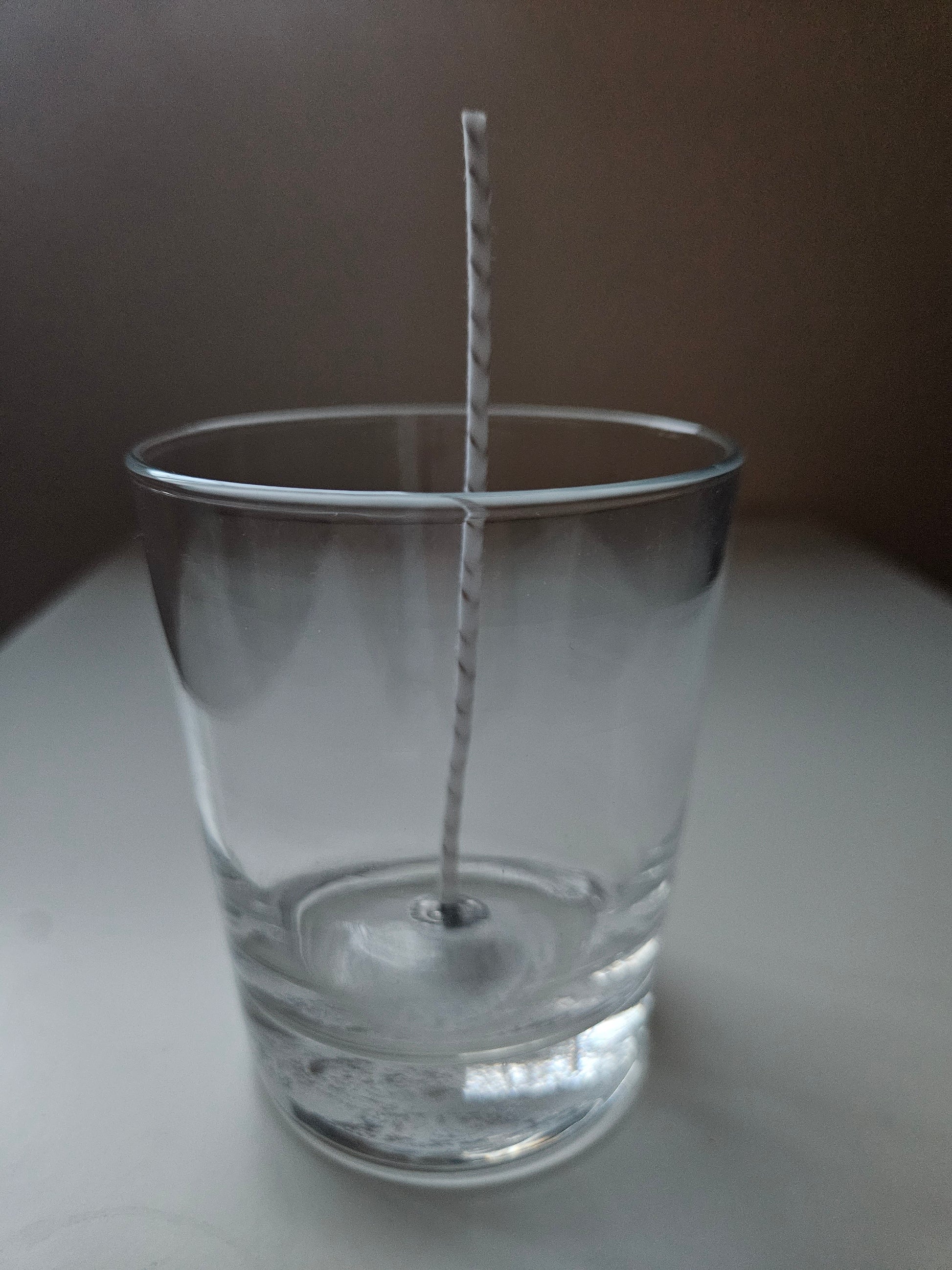 Leeres klares Glas mit weißem, gedrehtem Docht auf weißer Oberfläche vor dunklem Hintergrund